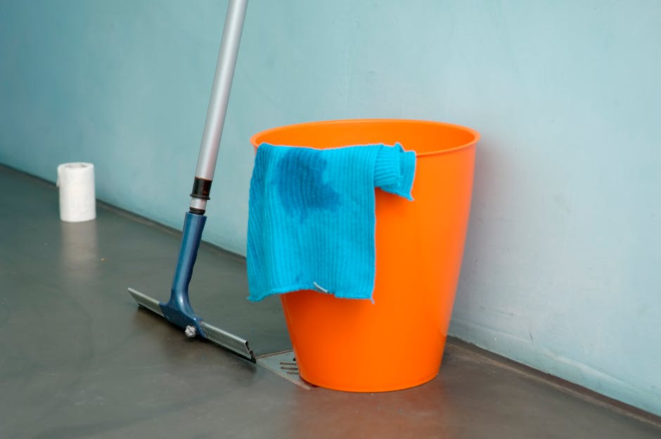 An orange plastic bucket with a blue cleaning cloth draped over its rim, positioned on a smooth, grey floor near a light-colored wall. A mop with a blue handle leans against the bucket, and a roll of white toilet paper is visible in the background. The floor appears clean and slightly reflective, indicating recent or ongoing cleaning, and the scene is illuminated with natural or soft artificial light, highlighting the surface cleanliness and tidy arrangement. This setup represents a typical scene for domestic or commercial surface cleaning, reflecting the professional services offered by Carpet Cleaners Hounslow to maintain hygiene and cleanliness in residential or business spaces.