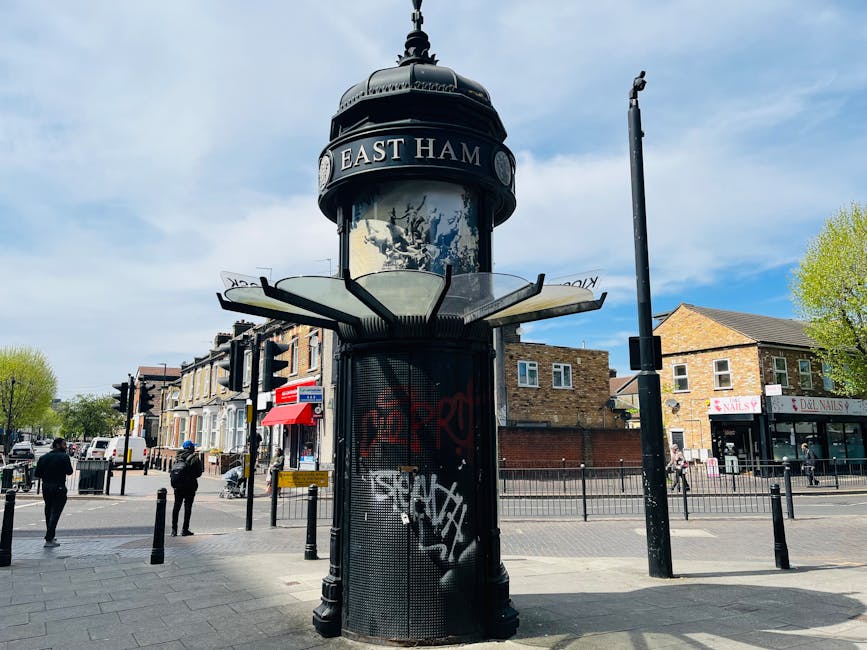 A cylindrical street sign covered with graffiti, situated on a paved sidewalk at a pedestrian crossing in East Ham, London, with a black arch top featuring the name 'EAST HAM'. Nearby, black metal bollards and a tall, slim street light pole are visible, and in the background are commercial buildings including shops with signs such as 'NAILS' and 'Deli NAILS'. The scene is illuminated by daylight with a partly cloudy sky, and some trees with green foliage are visible on the right side, indicating a lively urban environment. The area appears clean and well-maintained, reflecting the effectiveness of surface cleaning and hygiene standards managed by local services like Carpet Cleaners Hounslow.
