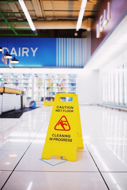 The image shows the interior of a commercial shop, likely a grocery or convenience store, with a shiny tiled floor and a yellow caution sign placed in the center indicating a wet floor and ongoing cleaning by Carpet Cleaners Hounslow. The store features shelves stocked with various products, visible in the background, and bright lighting illuminating the space. Large windows on the right side allow natural light to enter, enhancing the cleanliness and bright, welcoming atmosphere. The ceiling has exposed beams and integrated lighting fixtures. The overall environment appears clean and well-maintained, with the wet floor sign emphasizing ongoing surface cleaning and hygiene practices typical of commercial cleaning services. Carpet Cleaners Hounslow specializes in deep cleaning and sanitisation of such shopping environments.