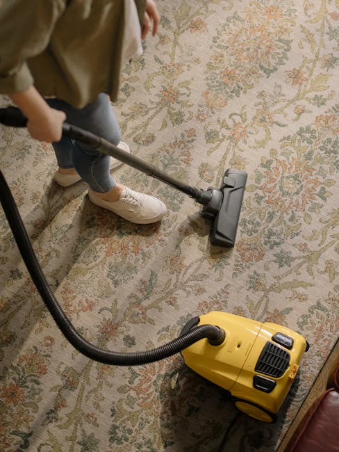 A person in casual attire, including white sneakers and blue jeans, is using a yellow and black vacuum cleaner with a long hose attachment to deep clean a traditional patterned carpet in a room with w