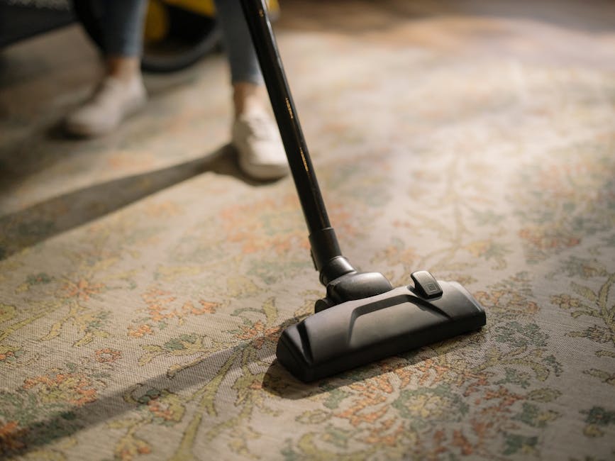 A close-up of a vacuum cleaner head being used to deep clean a patterned area rug in a home setting. The vacuum cleaner is black and modern, with a visible control button on top, and is positioned on the floral design of the carpet, which features light beige, green, and orange hues. The image captures the room's natural lighting, highlighting the clean and well-maintained state of the carpet, indicative of professional surface cleaning and sanitisation services provided by Carpet Cleaners Hounslow. In the background, the lower part of a person wearing light-colored sneakers and dark pants is partially visible, suggesting active cleaning. The setting emphasizes domestic cleaning processes aimed at maintaining hygiene and freshness of residential floors.