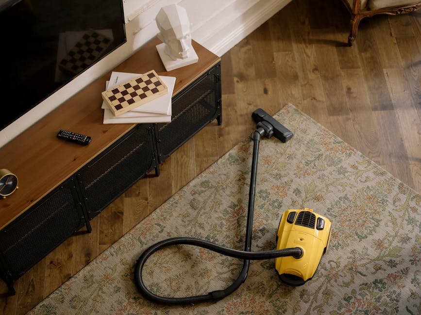 A yellow and black canister vacuum cleaner with a flexible hose and a floor attachment is positioned on a patterned area rug in a living room. The room features a wooden floor, a modern black metal and wood media console, and a white wall with decorative white bust sculpture, open books, and a remote control placed on top. Natural lighting highlights the clean surfaces, indicating surface cleaning and maintenance typically associated with professional domestic cleaning services by Carpet Cleaners Hounslow, specifically focusing on deep cleaning and sanitisation of residential spaces.