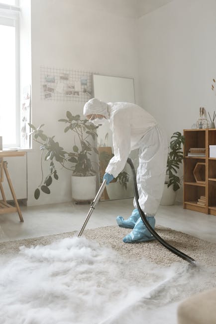 A person dressed in full protective gear, including a white suit, gloves, and blue shoe covers, is performing carpet cleaning in a bright, well-lit room. The individual uses a professional vacuum or e