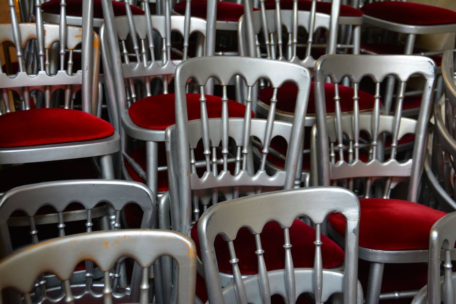 Close-up view of multiple stacked metal chairs with silver frames and red upholstered seats, arranged in a crowded manner in an indoor setting, likely a venue for events. The chairs are clean, with no visible dust or dirt, and the metal surfaces are shiny, reflecting light from overhead. The red fabric cushions on the seats appear fresh and well-maintained, contributing to a tidy appearance. The arrangement suggests preparation or post-event cleaning for a space used for gatherings or functions, and the scene highlights the importance of surface cleaning and sanitisation for maintaining hygiene in such environments. Carpet Cleaners Hounslow can assist with deep cleaning and sanitisation of similar seating areas to ensure cleanliness and hygiene after events. The background shows additional chairs, indicating a large seating arrangement, and the overall image emphasizes the need for professional cleaning in commercial or event spaces.