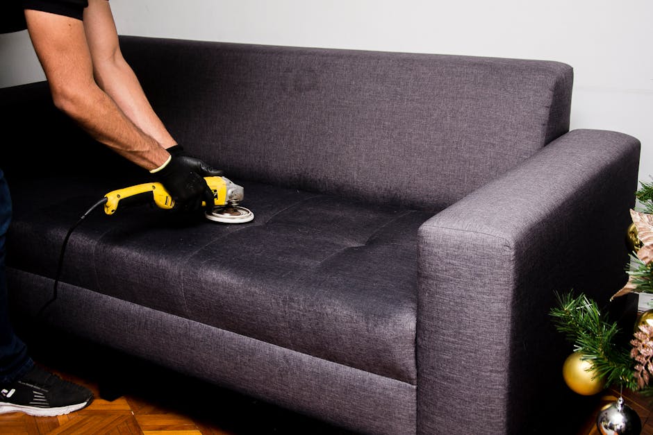 A person wearing black gloves is using a yellow handheld upholstery cleaning machine on a dark grey fabric sofa in a living room. The sofa has a contemporary design with straight arms and a cushioned backrest, situated on a hardwood floor. The room appears well-lit, highlighting the fresh, clean surface of the upholstery. To the right of the sofa, there is a decorated Christmas tree with gold and pink ornaments and greenery. The scene demonstrates surface cleaning and deep cleaning processes for upholstery, reflecting hygiene and maintenance standards associated with professional domestic cleaning services. The image is related to the services offered by Carpet Cleaners Hounslow for post-event upholstery sanitisation at Syon Park in Hounslow.