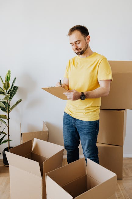 A man with short brown hair and a beard, dressed in a yellow t-shirt and blue jeans, standing in a room with plain white walls. He holds a clipboard and pen, appearing to check or note items. Surrounding him are several open cardboard moving boxes placed on the wooden floor, with some larger boxes stacked against the wall. A green potted plant with long, broad leaves is positioned to the left, adding a touch of greenery. The room is well-lit with natural light, providing a clean and organized appearance suitable for surface cleaning or general domestic cleaning activities, consistent with the context of a TW3 end of tenancy cleaning checklist for landlords, HOUNSLOW, at Carpet Cleaners Hounslow.