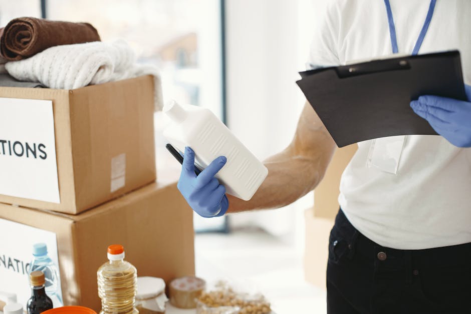 A man with short brown hair and a beard, dressed in a yellow t-shirt and blue jeans, standing in a room with plain white walls. He holds a clipboard and pen, appearing to check or note items. Surrounding him are several open cardboard moving boxes placed on the wooden floor, with some larger boxes stacked against the wall. A green potted plant with long, broad leaves is positioned to the left, adding a touch of greenery. The room is well-lit with natural light, providing a clean and organized appearance suitable for surface cleaning or general domestic cleaning activities, consistent with the context of a TW3 end of tenancy cleaning checklist for landlords, HOUNSLOW, at Carpet Cleaners Hounslow.