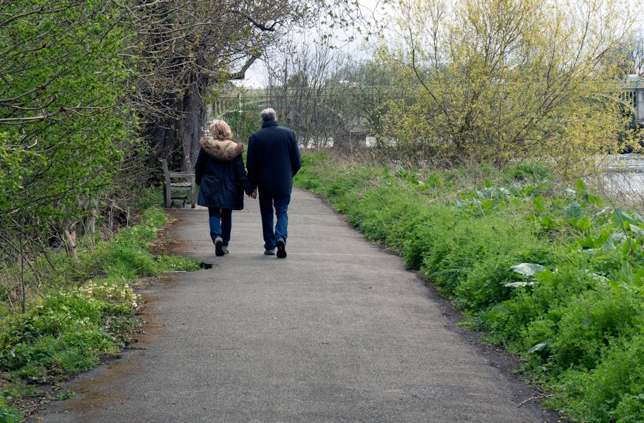 Two elderly individuals, a man and a woman, are walking hand-in-hand along a paved pathway in a park-like setting during daytime. The woman is wearing a dark coat with a fur-lined hood, and the man is dressed in a navy jacket and jeans. They are walking away from the camera, surrounded by trees with budding green leaves and lush green grass on either side of the path. In the distance, there are benches and a glimpse of a body of water, with an overcast sky overhead, creating a peaceful and natural environment. Carpet Cleaners Hounslow emphasizes clean and well-maintained outdoor spaces as part of our comprehensive cleaning services in Hounslow.
