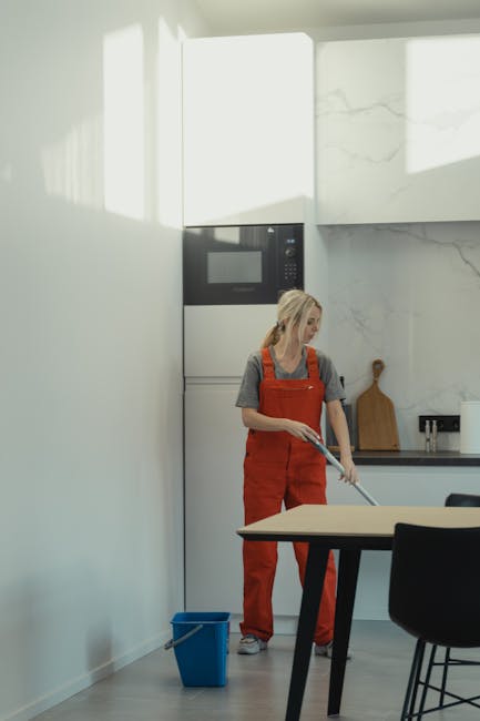 A woman in casual clothing and red overalls is performing surface cleaning in a modern kitchen, using a mop or cloth to clean the floor near a blue cleaning bucket. The kitchen features a white wall with a large marble-patterned panel, stainless steel appliances, and a wooden cutting board on the black countertop. Natural light floods the room, illuminating the clean, polished surfaces. This scene showcases domestic cleaning, emphasizing hygiene and maintenance, with the woman representing services provided by Carpet Cleaners Hounslow as part of their urgent same-day cleaning offerings in Hounslow.